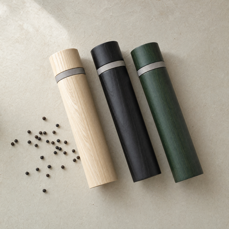 Three column pepper mills lying on their sides in ash, black ash and pine green, with scattered peppercorns